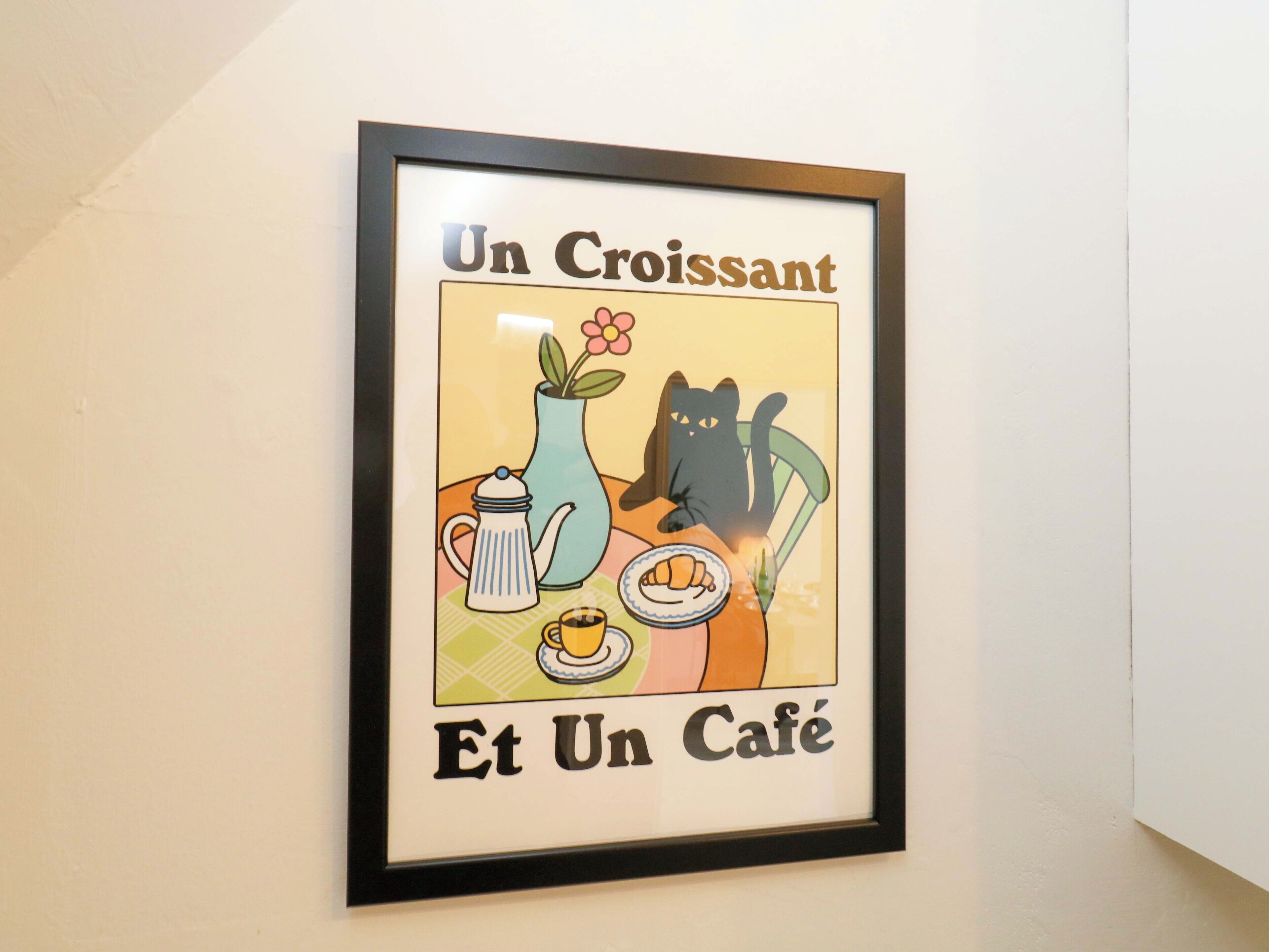 Un cuadro con un gato, una tetera, una flor y un croissant.