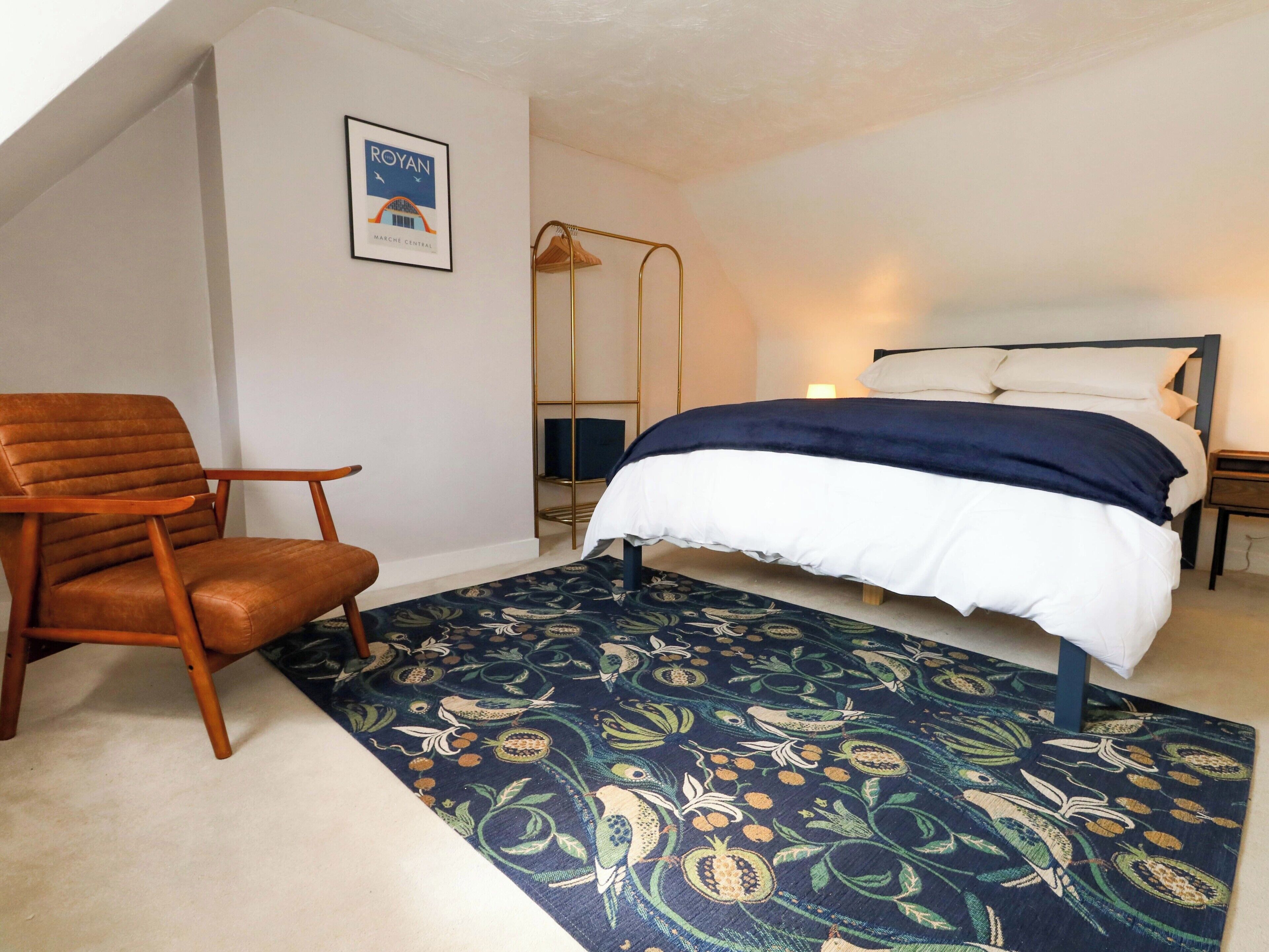 A bedroom with a bed, a chair, a patterned rug, and a framed picture on the wall.