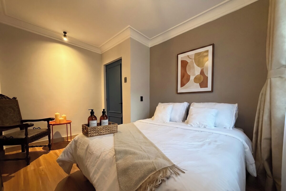 A hotel room with a bed, a chair, a small table with candles, and a wall-mounted artwork.