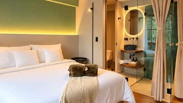 Une chambre moderne avec un grand lit, une salle de bain fermée par des parois de verre et une vue sur la verdure environnante.