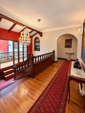 A grand staircase with a wooden railing, a chandelier, and a red carpet leading to an arched doorway. - Sereno Hostels (Bogota)