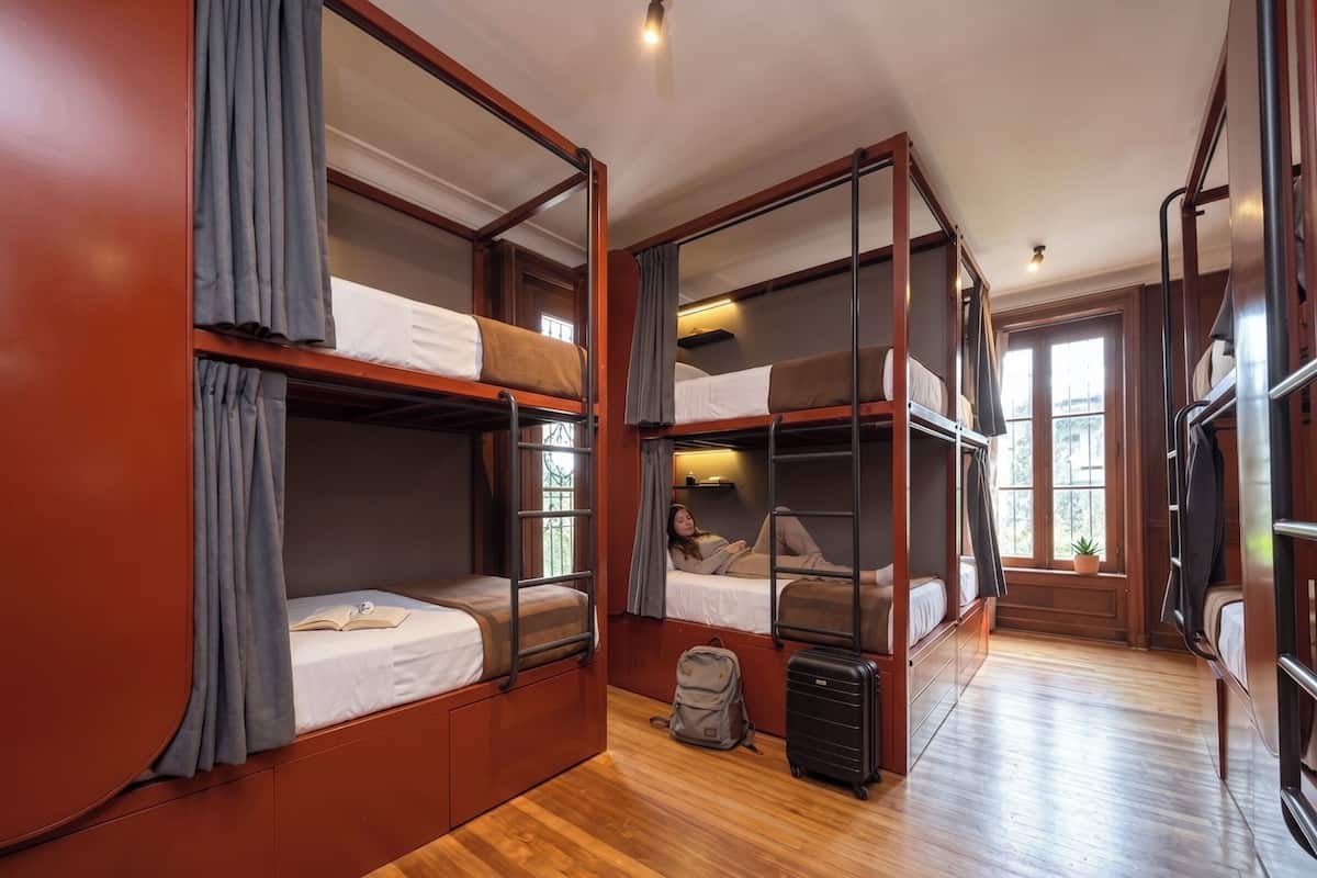A room with bunk beds, wooden flooring, a suitcase, and a window with a view.