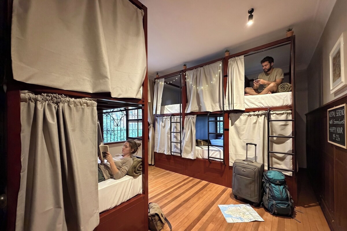 A dormitory-style room with bunk beds, a wooden floor, and a window with curtains.