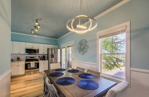 A kitchen with white cabinets, a stainless steel refrigerator, and a dining area with a wooden table and blue placemats. - Coral Cabana: Salt Air and Sunset Views! (Surf City)