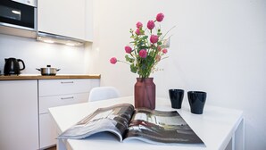 Eine Küche mit weißen Schränken, einem schwarzen Wasserkocher, einer braunen Vase mit rosa Blumen und zwei schwarzen Tassen auf einem Tisch.