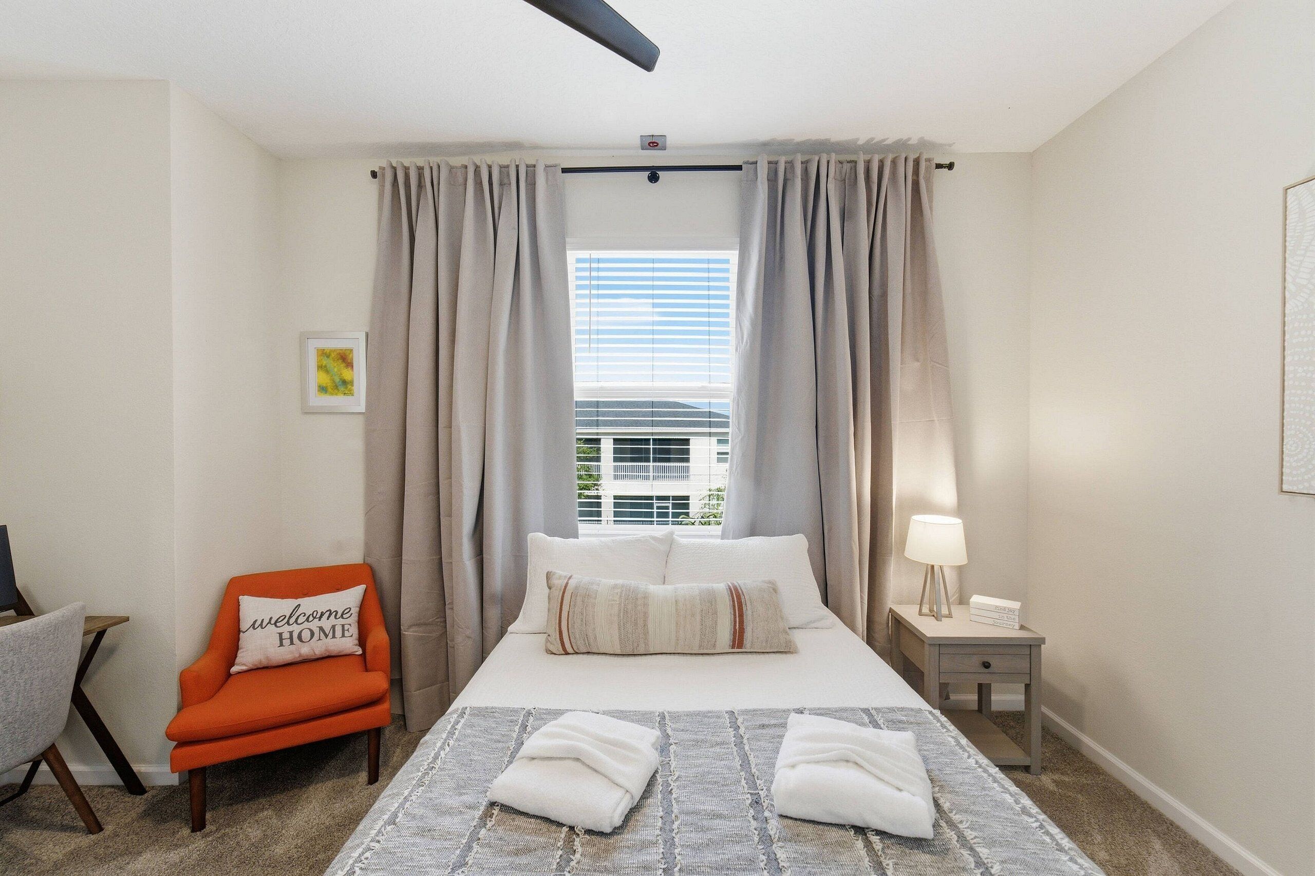 A hotel room with a bed, a chair, a nightstand, a window with curtains, and a ceiling fan.