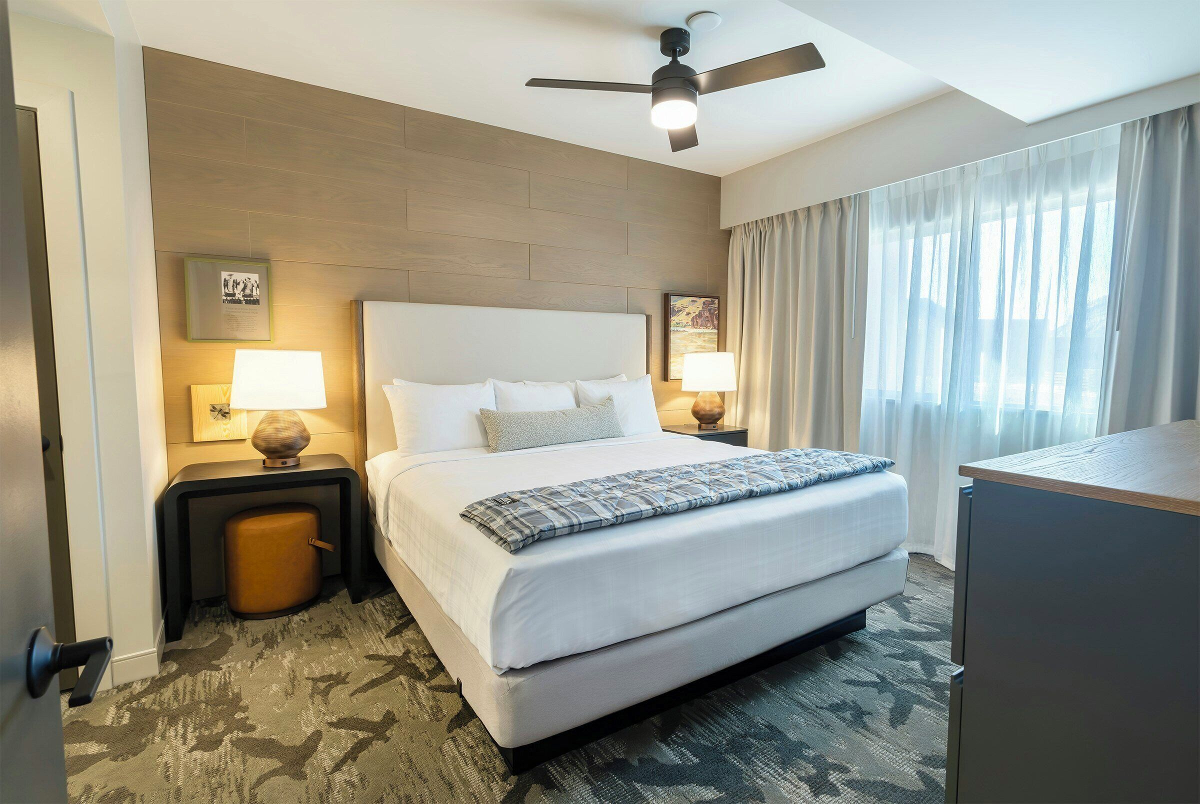 A hotel room with a large bed, two bedside tables with lamps, a ceiling fan, and a view of the outdoors through the window.