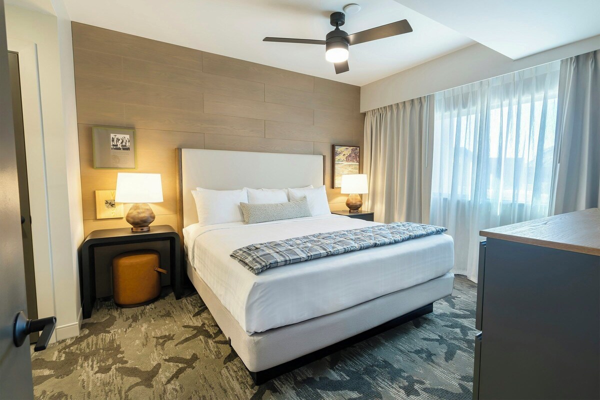 A hotel room with a large bed, two bedside tables with lamps, a ceiling fan, and a view of the outdoors through the window.