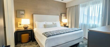 A hotel room with a large bed, two bedside tables with lamps, a ceiling fan, and a view of the outdoors through the window.