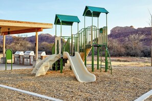 A playground with slides and climbing structures, surrounded by a gravel area and a barbecue setup. - EBAC Moab (Moab)
