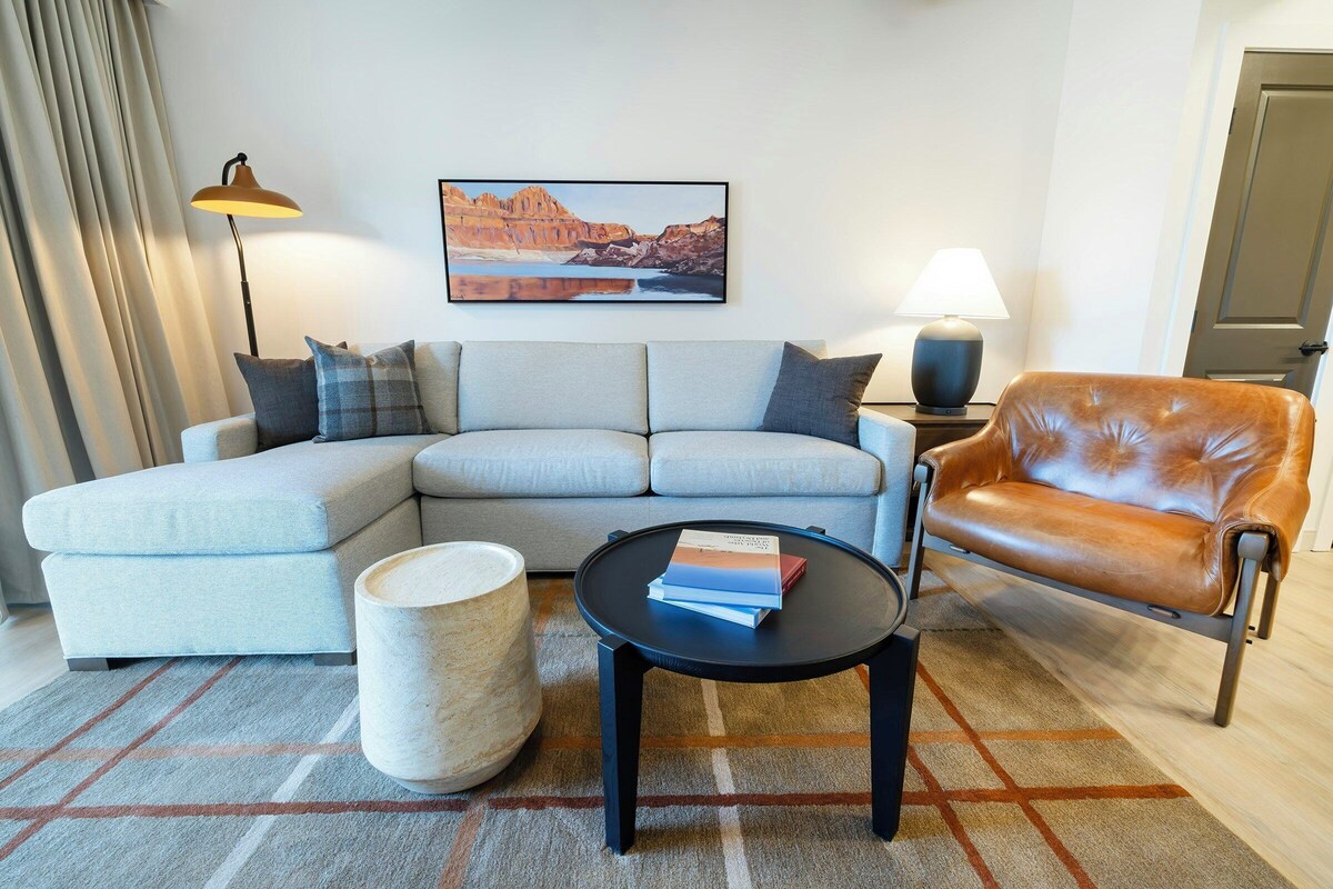 A living room with a sectional sofa, a leather armchair, a round coffee table, a lamp, and a painting on the wall.