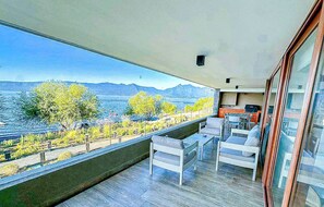A balcony with a view of a lake and mountains, furnished with white chairs and a table.