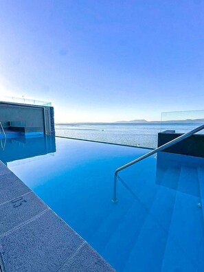 A clear blue infinity pool with a metal railing, overlooking a calm sea and distant land.