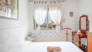 Ein Schlafzimmer mit Bett, einem Fenster mit Vorhängen, einem Holzstuhl, einem kleinen Tisch mit Krug und einem Spiegel.