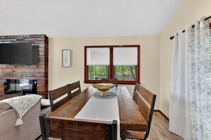 A dining area with a wooden table, chairs, a decorative bowl, a television on a brick wall, and a window with blinds.
