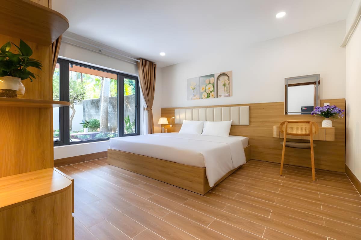 A modern bedroom with a large bed, wooden headboard, a bedside table, a chair, a window with curtains, and a wall-mounted mirror.