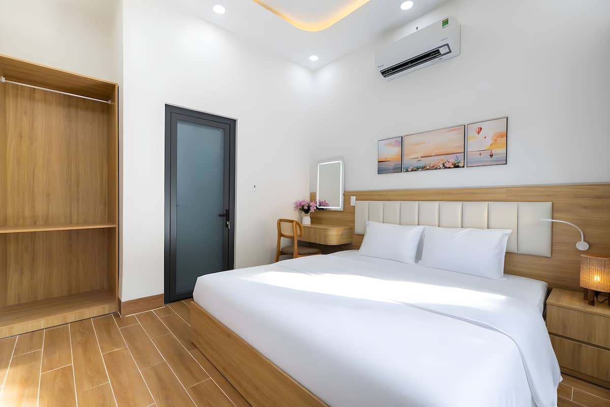 A hotel room with a large bed, a wooden wardrobe, a desk with a chair, a bedside table with a lamp, and wall-mounted artwork.