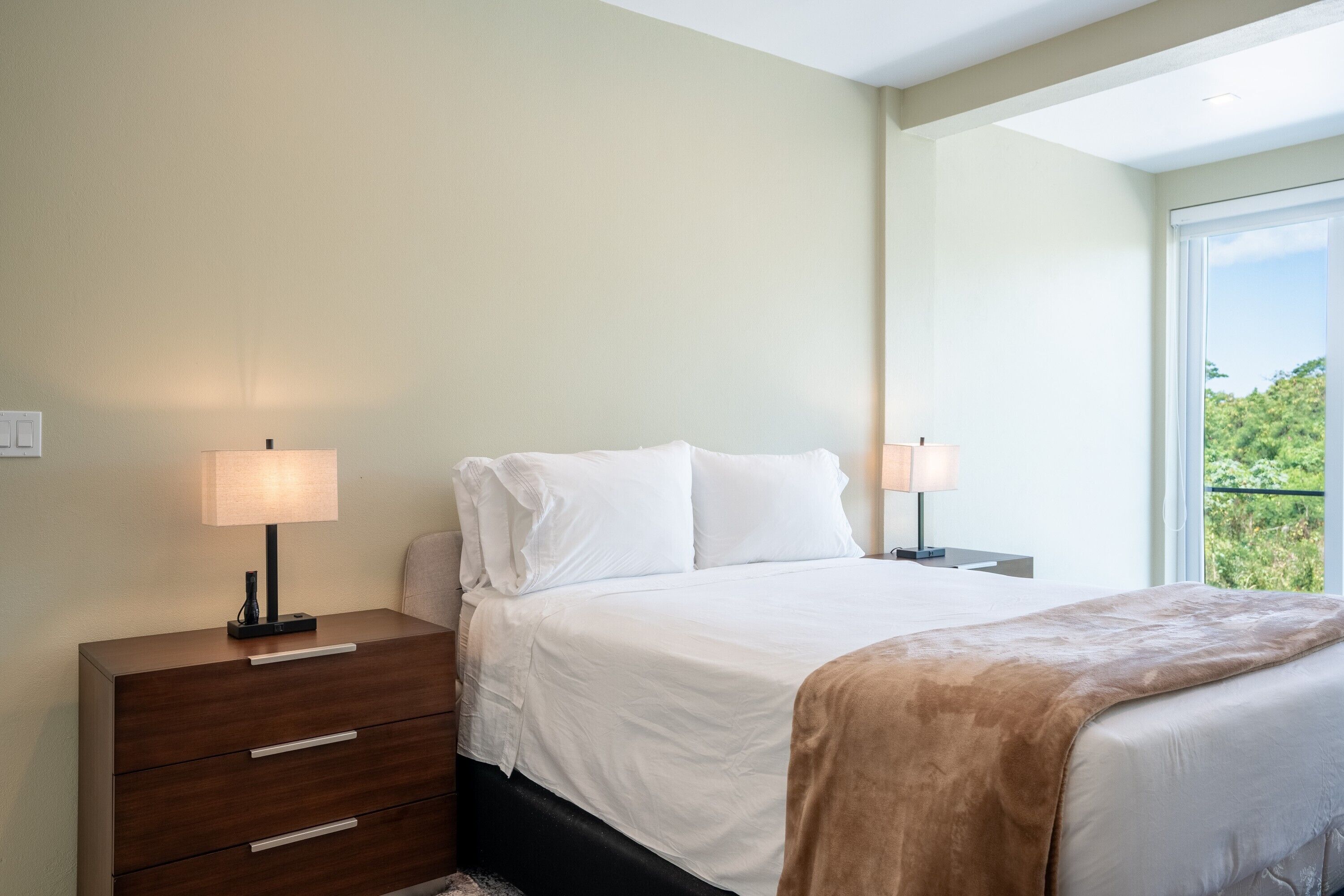 A bedroom with a bed, two bedside tables, a nightstand, and a window with a view.