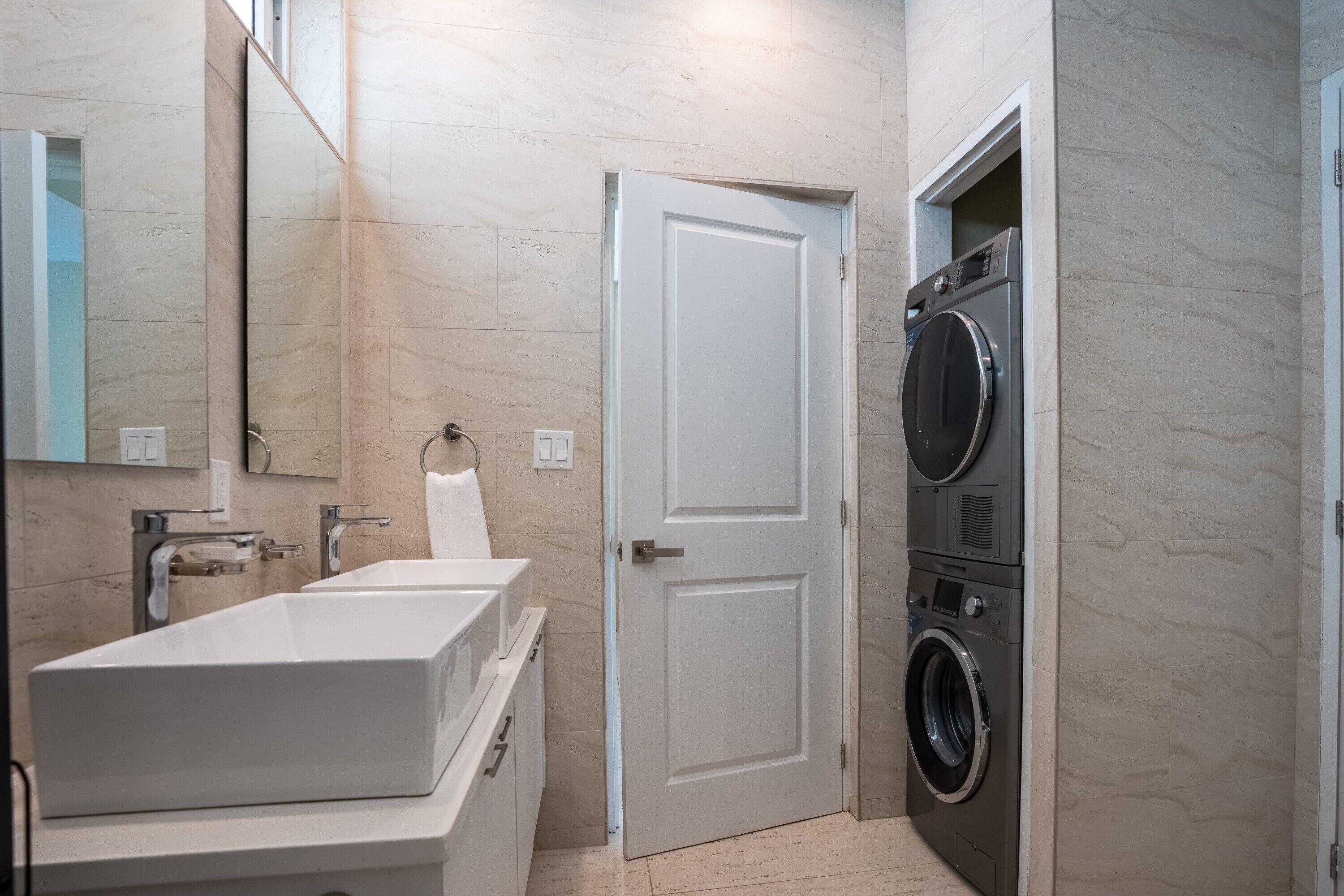 A bathroom with a white sink, a mirror, a towel dispenser, and a door leading to a laundry area with a washing machine.