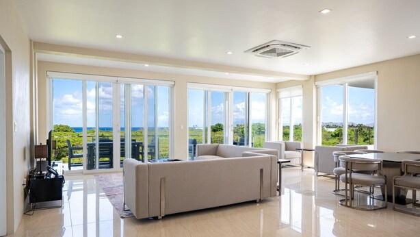 A spacious living area with a large sofa, a dining table, and chairs, with a view of greenery and a distant landscape through large windows. - Belmont Villas (Vaughans)
