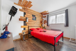 Quarto com cama, painéis de parede em madeira, janela com cortinas, mesa pequena com abajur e uma planta.