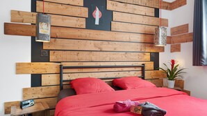 Une chambre avec un lit, une tête de lit en bois, deux lampes fixées au mur, une petite table avec une plante et une couverture de lit rouge.