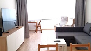 Una sala de estar moderna con un televisor de pantalla plana, un sofá, una mesa de comedor con sillas y un gran ventanal con cortinas.