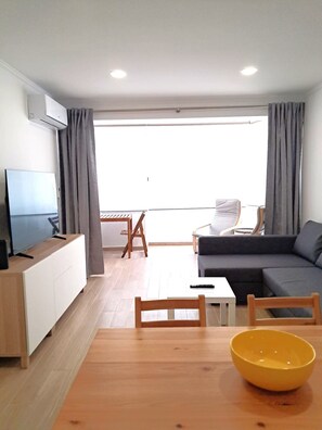 Ein modernes Wohnzimmer mit Flachbildfernseher, einer Couch, einem Esstisch mit Stühlen und einem großen Fenster mit Vorhängen.