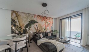 Ein modernes Wohnzimmer mit einer großflächigen Wandmalerei im tropischen Stil, einem Glastisch mit Stühlen, einer Couch mit Kissen und einem kleinen Beistelltisch.