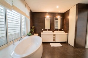 A modern bathroom with a large bathtub, two sinks, dark tiled walls, and a window with blinds.