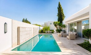 A modern outdoor pool area with a clear blue pool, surrounded by a white wall and a patio with seating.