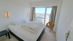 A modern bedroom with a large bed, a chair, a nightstand, and a view of the city through large windows. - Sea View Family Stay for 8 (Koksijde)