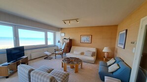 A living room with a television, two sofas, a coffee table, and a chair. There are large windows with a view of the beach. - Sea View Family Stay for 8 (Koksijde)