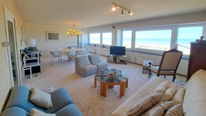 A spacious living area with a large window overlooking the beach, a dining table, a sofa, and a television. - Sea View Family Stay for 8 (Koksijde)