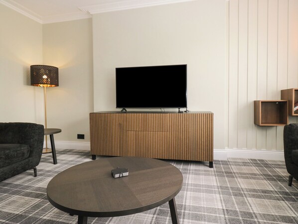 A living room with a flat-screen TV on a wooden cabinet, a round coffee table, a sofa, and a floor lamp. - 5 Thornbarrow Road (Windermere)