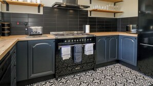 A modern kitchen with a stove, oven, and black cabinets. There are wooden shelves above the counter holding cups and glasses. - 5 Thornbarrow Road (Windermere)