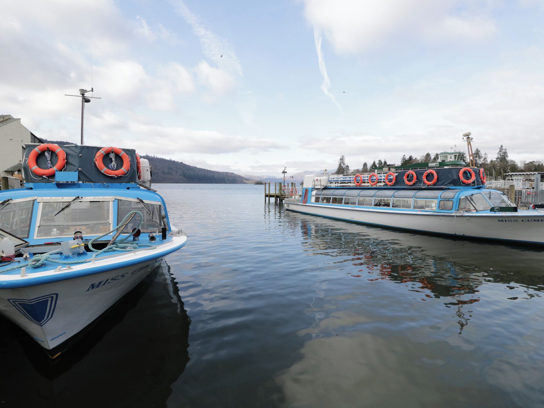 Two boats docked at a marina with lifebuoys on the side, calm water, and a cloudy sky.