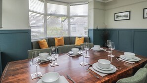 A dining area with a wooden table set for six, a green upholstered bench, and a bay window with blinds. - 5 Thornbarrow Road (Windermere)