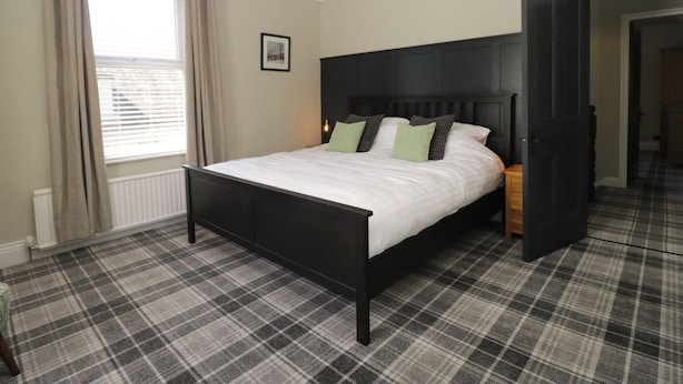 A bedroom with a bed, a headboard, a window with curtains, a radiator, and a plaid carpet. - 5 Thornbarrow Road (Windermere)