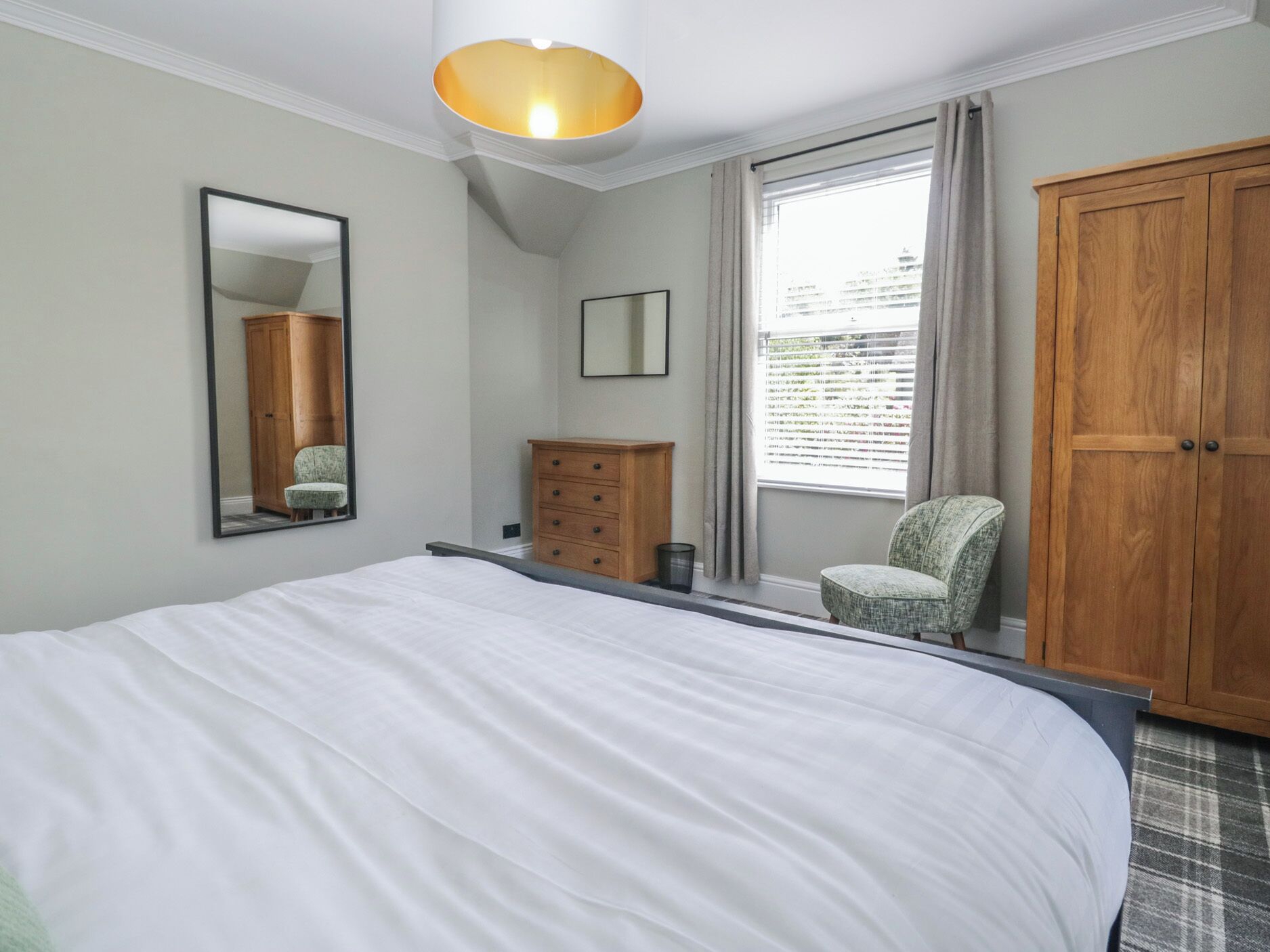 A bedroom with a large bed, a wooden wardrobe, a small chair, a mirror, and a window with curtains.