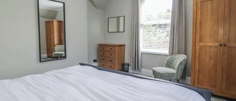 A bedroom with a large bed, a wooden wardrobe, a small chair, a mirror, and a window with curtains.