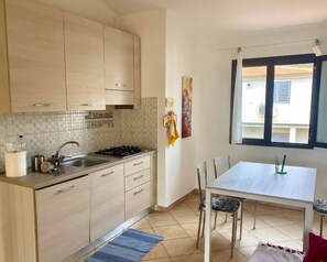 A kitchen with a sink, stove, and cabinets, a dining table with chairs, and a window with curtains. - SA MARINA HOUSE #3. Cozy house for families with children, youth and animal (Sassari)
