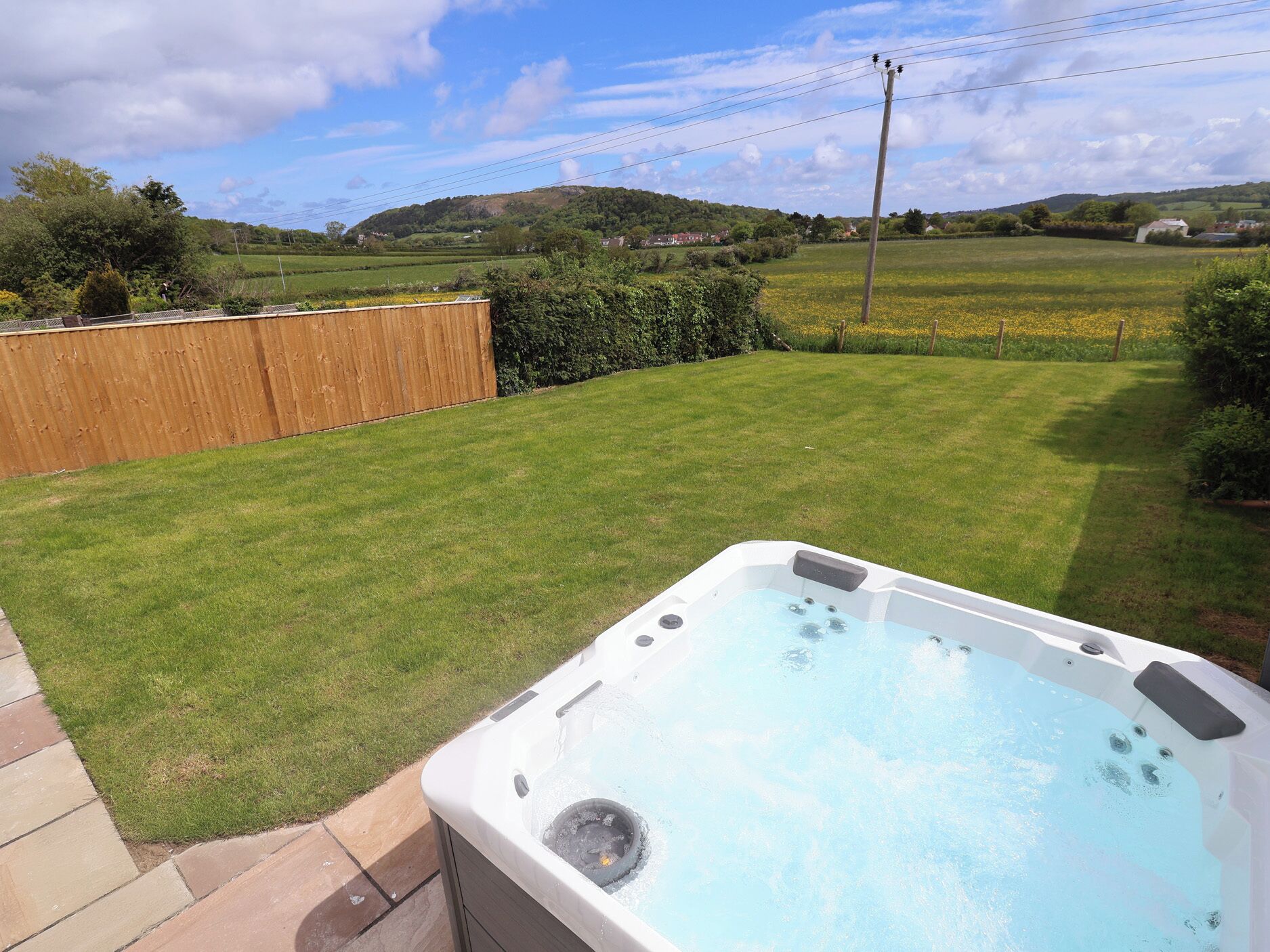 A hot tub in a backyard with a wooden fence, a grassy lawn, and a hilly landscape in the background.