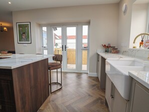 A modern kitchen with a central island, a dining area with a chair, and a sliding glass door leading to a balcony. - Bella Vista (Llandudno)