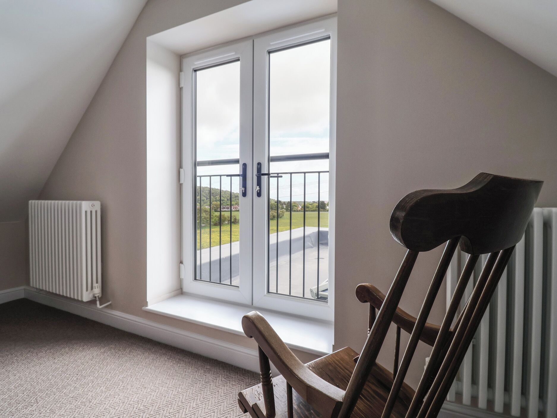 Una habitación con techo inclinado, un radiador, una ventana con vistas al exterior y una silla de madera.