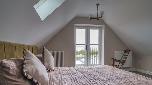 A bedroom with a sloped ceiling, a bed with a quilted cover, two pillows, a wooden chair, and a radiator. - Bella Vista (Llandudno)