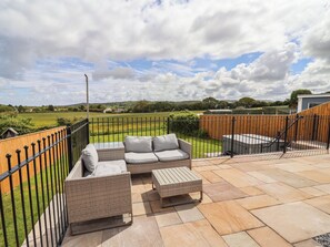 An outdoor patio with wicker furniture, a wooden fence, and a view of a green field. - Bella Vista (Llandudno)
