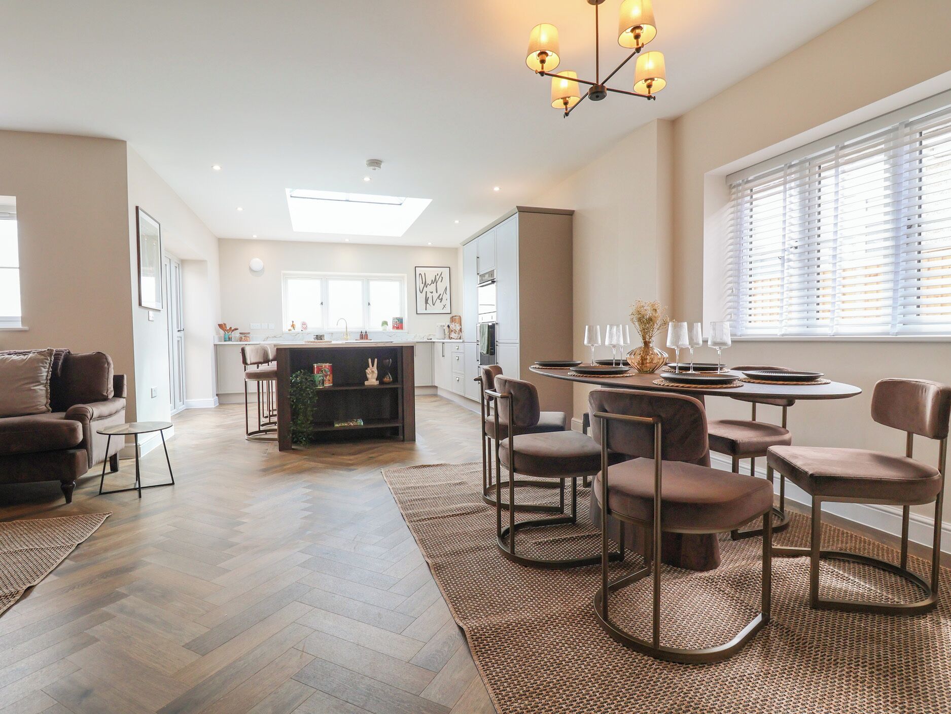 A spacious living area with a dining set, a kitchen in the background, and a large window with blinds.