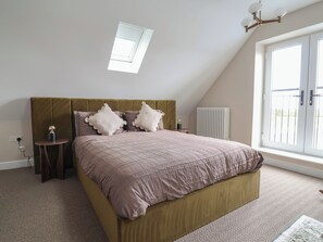 A bedroom with a sloped ceiling, a bed with a headboard, a bedside table with a lamp, and a window with a view. - Bella Vista (Llandudno)
