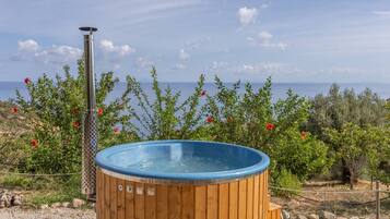 A hot tub with a wooden exterior and a metal heating element, surrounded by greenery and overlooking a body of water.